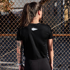A woman with a ponytail faces a chain-link fence, wearing a black GORUCK Women's Cropped Tee - Poly-Blend featuring a logo on the back. Her left arm showcases an impressive tattoo sleeve, with part of a building in the background.