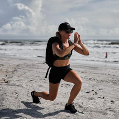 Woman rucking with GORUCK backpack performing lunges on sandy beach near ocean waves