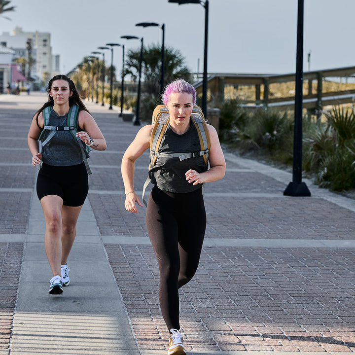 Two women jog alongside a beach, each carrying backpacks and confidently wearing GORUCK Women's Training Leggings - ToughFlex. Made for comfort and support, these squat-proof leggings suit their seaside run perfectly over the brick path.