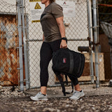 Wearing Women’s Training Leggings - ToughFlex, a person in athletic wear carries a black backpack. Confidently walking near an industrial area and chain-link fence, they are assured every step is squat-proof and stylish.