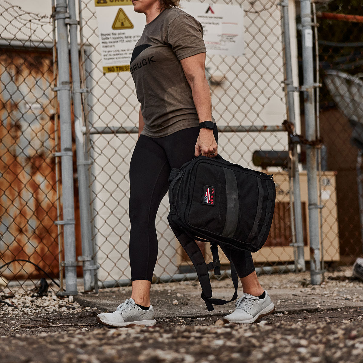 A person in athletic wear, wearing high-waisted Training Leggings and carrying a black backpack, walks outdoors near a fenced area with warning signs.