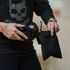A person with tattooed hands holds a black Packable Bullet Ruck - Ripstop ROBIC® and a folded item featuring a USA flag patch.