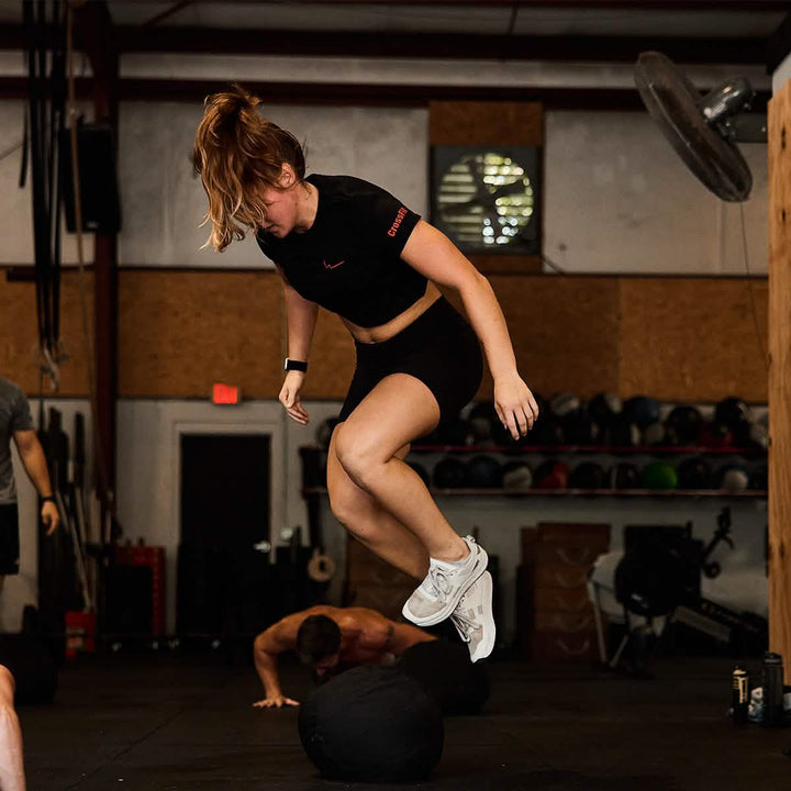 A person with brown hair in a ponytail jumps over a large exercise ball inside a gym. Wearing a Women's Subtle Spearhead x CrossFit Cropped Tee - Poly-Blend by GORUCK and shorts, paired with white sneakers, they effortlessly showcase their workout prowess. Other people are exercising in the background, surrounded by premium poly-blend gym equipment.
