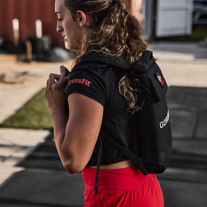 A person with long hair, wearing a Women's Subtle Spearhead x CrossFit Cropped Tee by GORUCK and red pants, stands outdoors holding a small black backpack. The sunny setting includes grass and concrete, making it an ideal space for a dynamic workout.