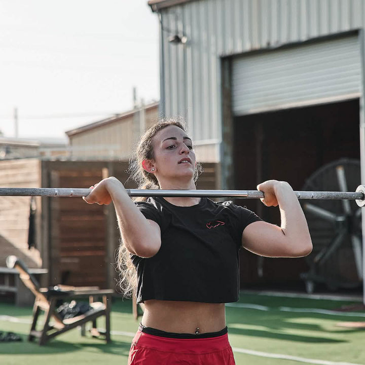 A woman is lifting a barbell outdoors in a gym setting while wearing the Women's Subtle Spearhead x CrossFit Cropped Tee - Poly-Blend by GORUCK, paired with red shorts. The area features artificial grass and wooden structures, with a large industrial fan in the background.