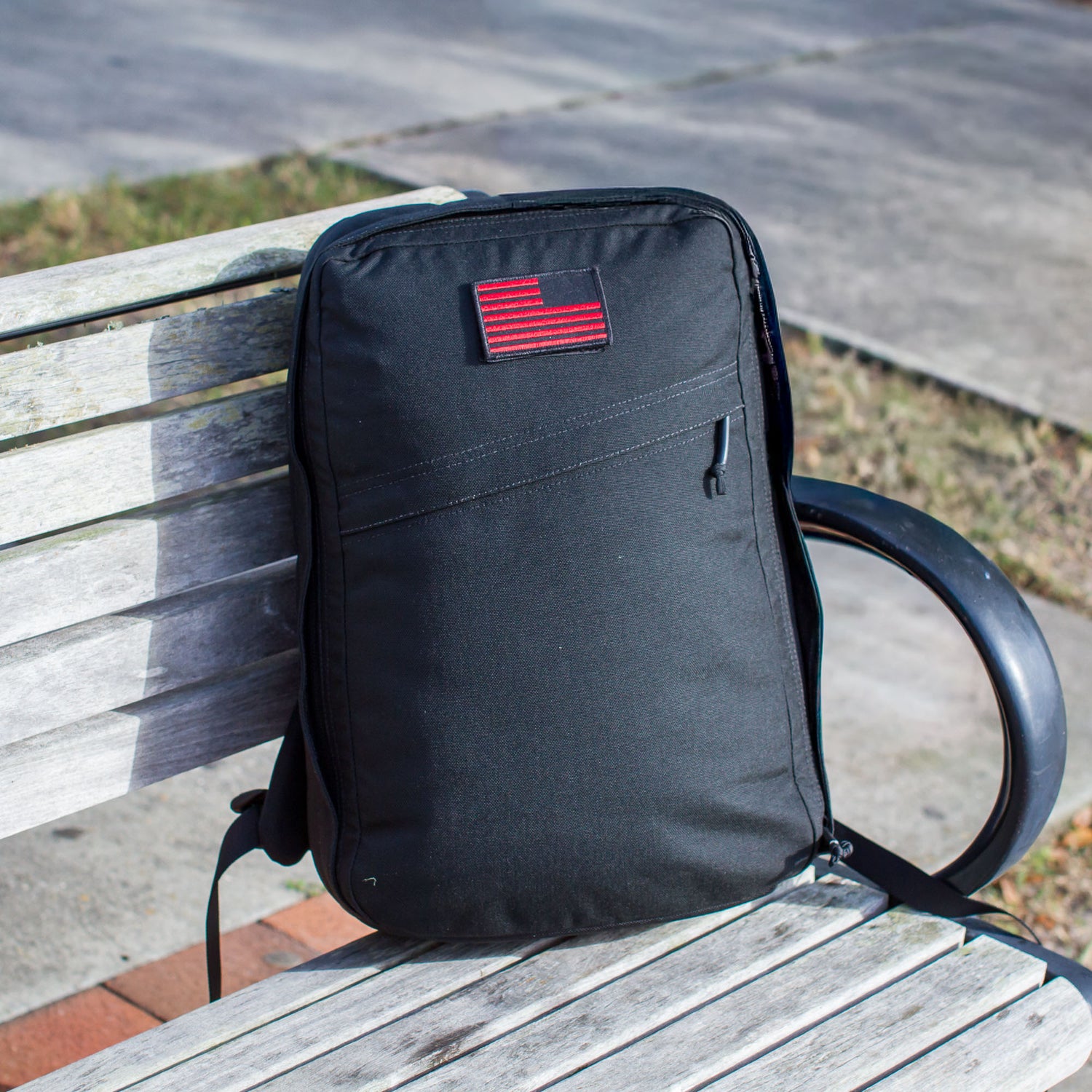 The Shadow Ruck, a lightweight backpack with a red patch, rests on a wooden bench outdoors—ideal for everyday carry.