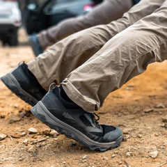 Two people wearing beige pants and black hiking shoes, featuring the GORUCK Merino Challenge Socks - Trail Cuff, sit on a rocky, dirt surface. The focus is on their legs and footwear. Cars are partially visible in the blurred background, adding an adventurous touch to the scene.