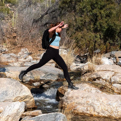 A person outfitted in a blue top, black leggings, a backpack, and durable rucking shoes is captured mid-jump over a rocky stream in a natural landscape with dry grass and trees, wearing GORUCK's Merino Challenge Socks - Trail Cuff.