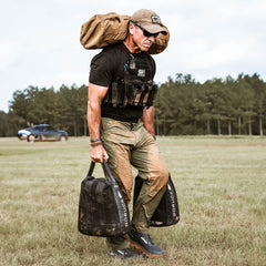 Clad in tactical gear, including a weighted vest and hat, an individual hauls large weighted bags across a grassy field. Their feet stay comfortable in GORUCK's Merino Challenge Socks - Trail Cuff paired with rucking shoes. A brown rucksack is secured to their back, while a blue SUV is parked near a line of trees in the background.