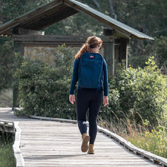 A woman with a backpack strolls along the boardwalk toward a small wooden structure, embraced by lush green foliage. Sporting a blue top and black leggings, she steps confidently in GORUCK Merino Challenge Socks - Trail Cuff boots, her hair tied in a ponytail.