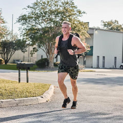 Man rucking outdoors with GORUCK backpack, wearing black tank and camo shorts, on sunny day