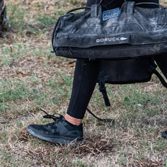 Person wearing black athletic shoes and leggings carrying a black GORUCK duffel bag outdoors on grass