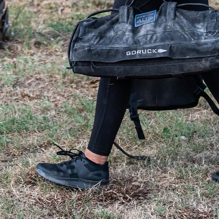 Clad in the Women's Rough Runner - Blackout black leggings and sneakers featuring an EVA midsole, a person carries a large black GORUCK bag over their shoulder while walking on grass. The bag displays visible GORUCK branding and boasts a rough, rugged look ideal for outdoor activities.