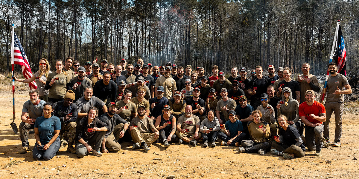A large group of people posing outdoors on dirt, many wearing athletic gear, with two American flags nearby.