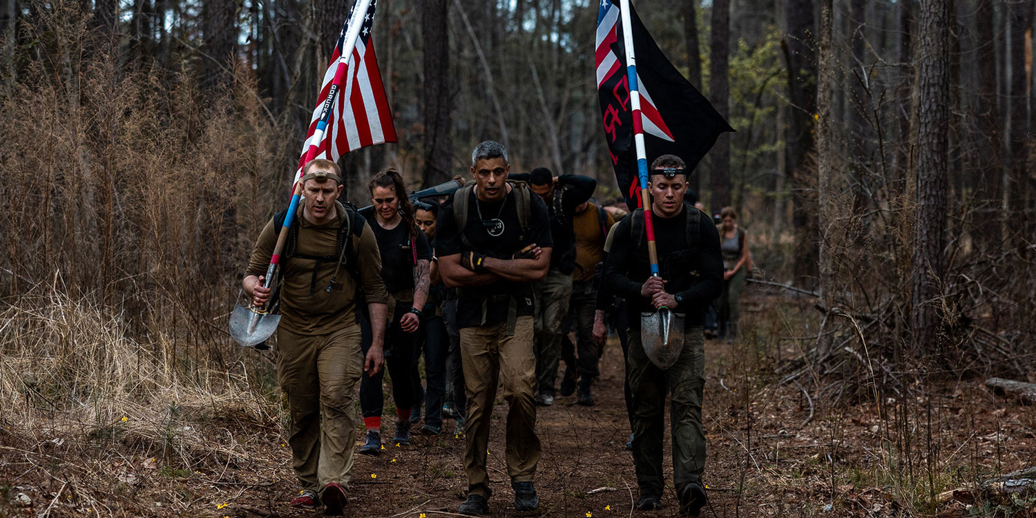 A group of people hiking through a forest, carrying flags and shovels, with serious expressions.