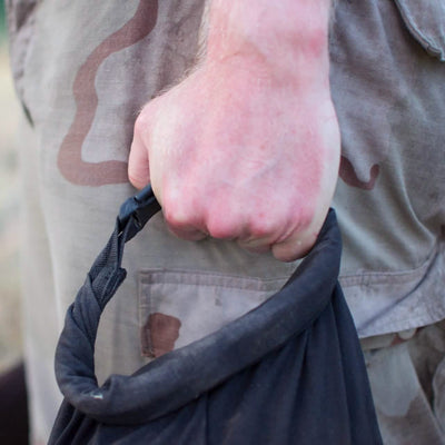 A person with light skin wearing camo pants is gripping a GORUCK Brick Bag (5-50 LBS) tightly, resembling military gear. The focus is on the hand and the texture of the fabric.