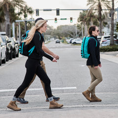 Three people with Bullet Ruck Classic - Cordura daypacks cross a street as palm trees sway gently in the background.