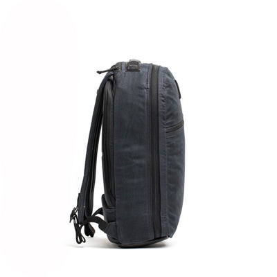 A side view of the Bullet Ruck - Waxed Canvas by GORUCK features a minimalist dark gray design with multiple zippers and adjustable shoulder straps, set against a plain white background. Its sleek and modern appeal is reminiscent of the classic retro canvas rucksack.