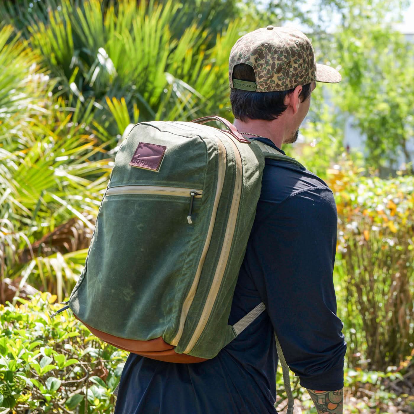 A man in a cap carries his Waxed Canvas 18L Bullet Ruck outdoors, its dual compartments securely holding his essentials as he strolls through lush greenery.