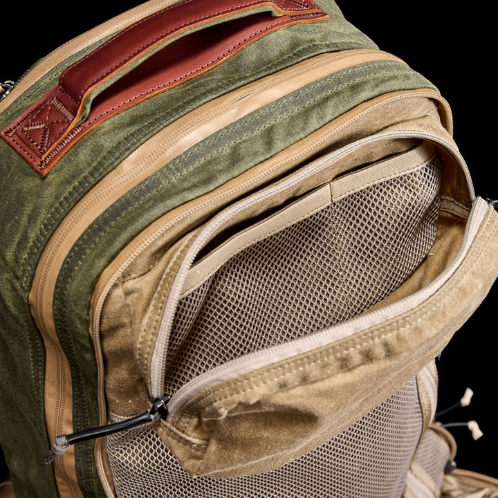 The green and tan Waxed Canvas 18L Bullet Ruck features a mesh front pocket, leather handle, and dual compartment design against a black backdrop. Built to last, it includes the renowned Scars Lifetime Guarantee.