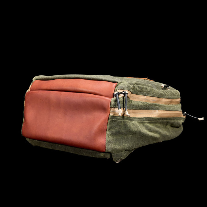 The Waxed Canvas 18L Bullet Ruck, featuring green and brown leather trim, is displayed on a black background with its visible zippers and pockets.