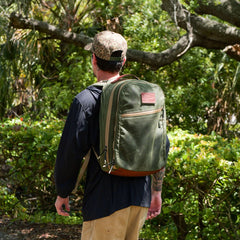 In the midst of lush greenery, someone wearing a camouflage cap walks with confidence, carrying a green Waxed Canvas 18L Bullet Ruck.