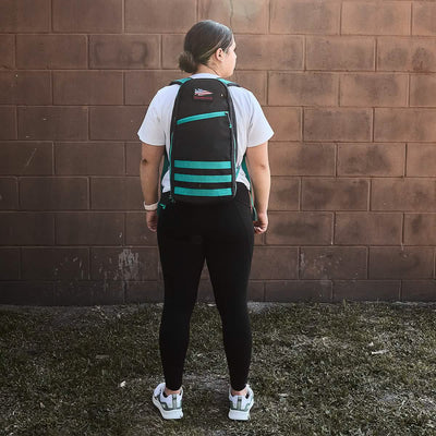 Standing on the grass with their back to the camera, a person wearing a white shirt and black leggings sports a teal and black GORUCK Bullet Ruck - 10L featuring padded straps. The durable Cordura fabric of the backpack enhances both style and functionality against the backdrop of a brown brick wall.