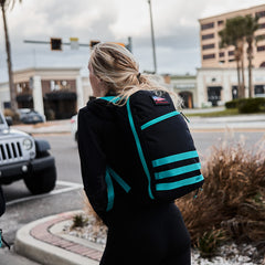 A person with blonde hair walks down the street, sporting a large black and teal Bullet Ruck Classic - Cordura backpack.