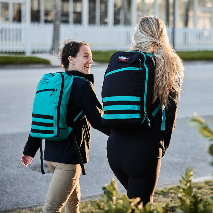 Two people walk outdoors with backpacks, smiling and talking near a white fence and lush greenery. Their Bullet Ruck Classic daypacks, crafted from robust Cordura, add a sense of adventure to the scene.