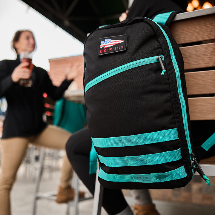 A close-up shows the Bullet Ruck Classic - Cordura, a black and teal 15L daypack, resting on a chair while two people chat and drink in the background.