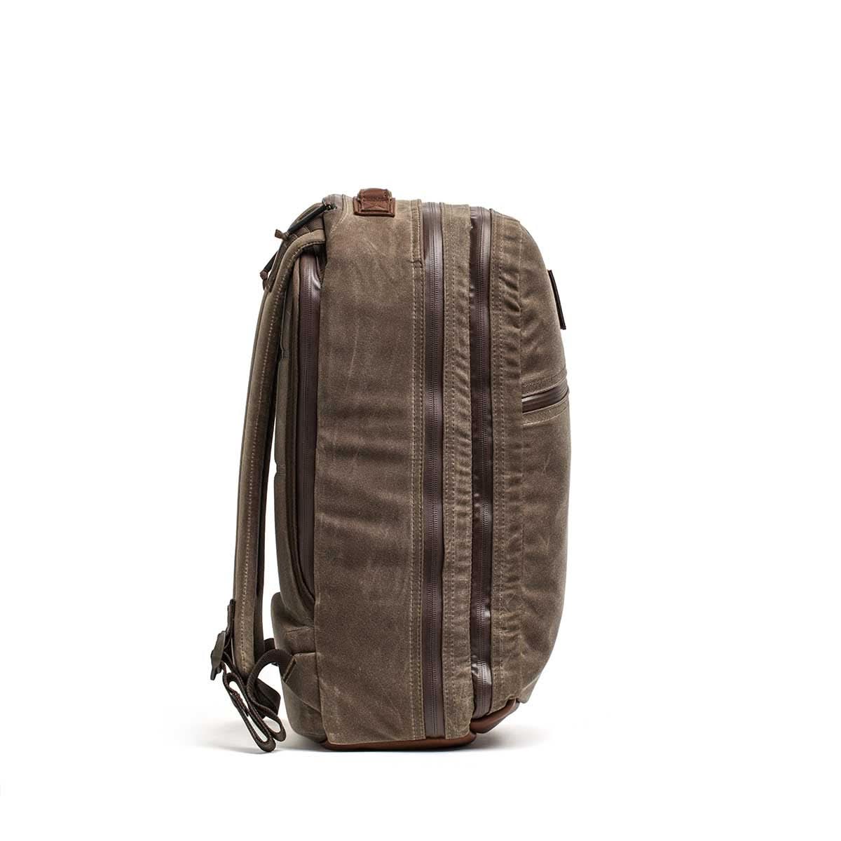 Side view of the Waxed Canvas 18L Bullet Ruck backpack in brown, featuring multiple zippers, dual compartments, and adjustable shoulder straps, set against a white background.