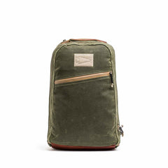 The Waxed Canvas 18L Bullet Ruck in green, with a brown base, offers dual compartments, a front zipper pocket, and a triangular logo. Its durable design is backed by the Scars Lifetime Guarantee for lasting quality and style.