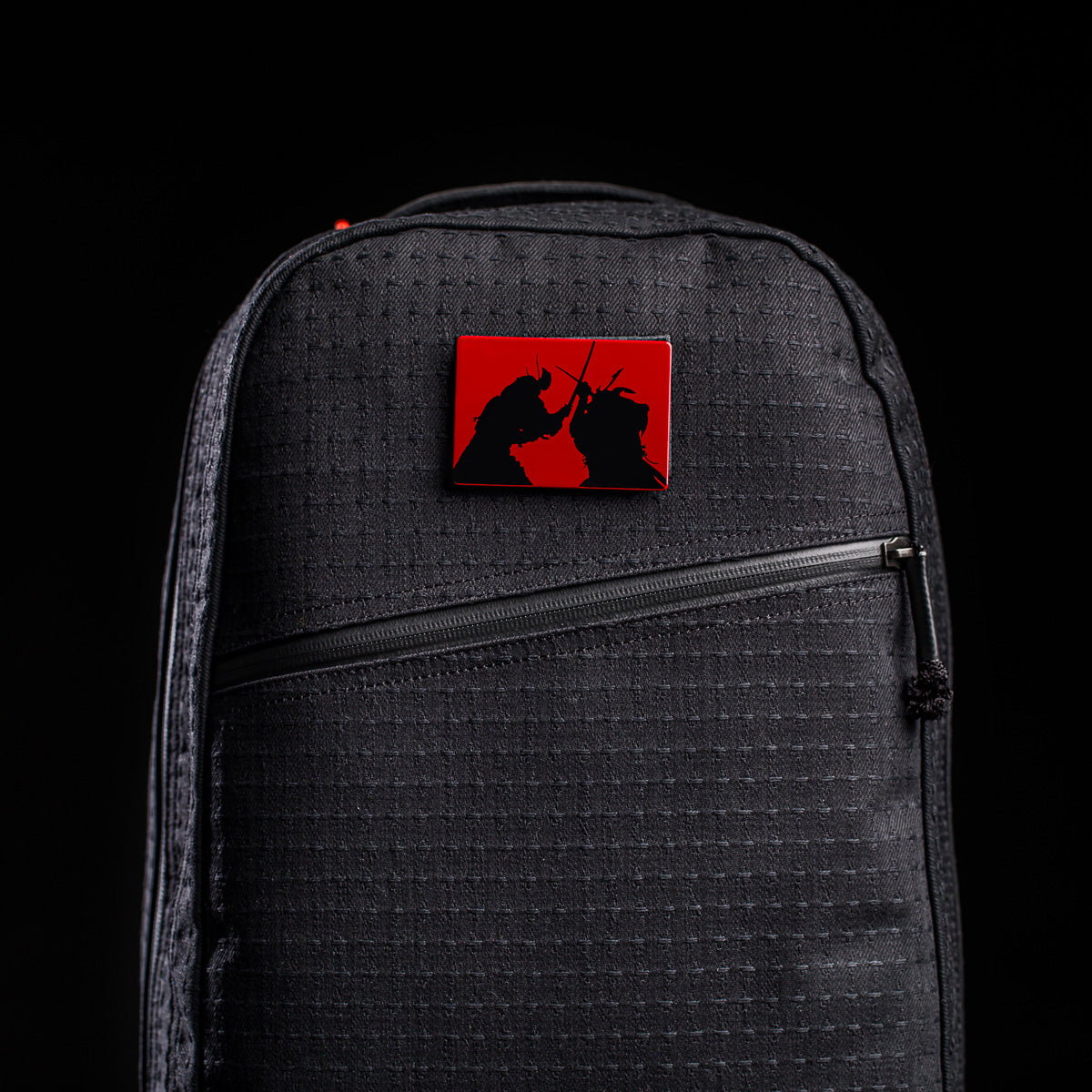 The limited-edition Bullet Ruck x Carryology - GRXC2 Samurai 2.0 is a black backpack featuring a red rectangular pin with two samurai silhouettes dueling—an homage to classic samurai showdowns.
