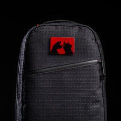 The limited-edition Bullet Ruck x Carryology - GRXC2 Samurai 2.0 is a black backpack featuring a red rectangular pin with two samurai silhouettes dueling—an homage to classic samurai showdowns.
