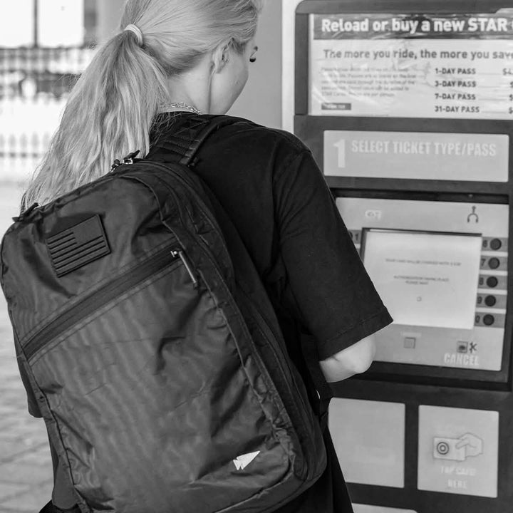 A person with long hair and a large Bullet Ruck Double Compartment - Dyneema by GORUCK stands in front of a ticket machine, selecting a ticket type. The machine displays various options, including passes for different durations. Black and white image.
