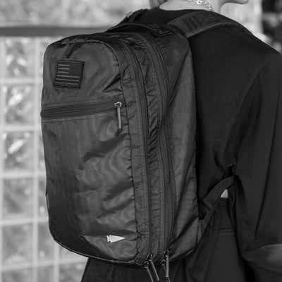 A person wearing a large black GORUCK Bullet Ruck Double Compartment - Dyneema, featuring multiple zippers and a patch on the back, stands against a wall of glass blocks. Dressed in dark clothing, they are prepared to travel hands-free with ease and style.