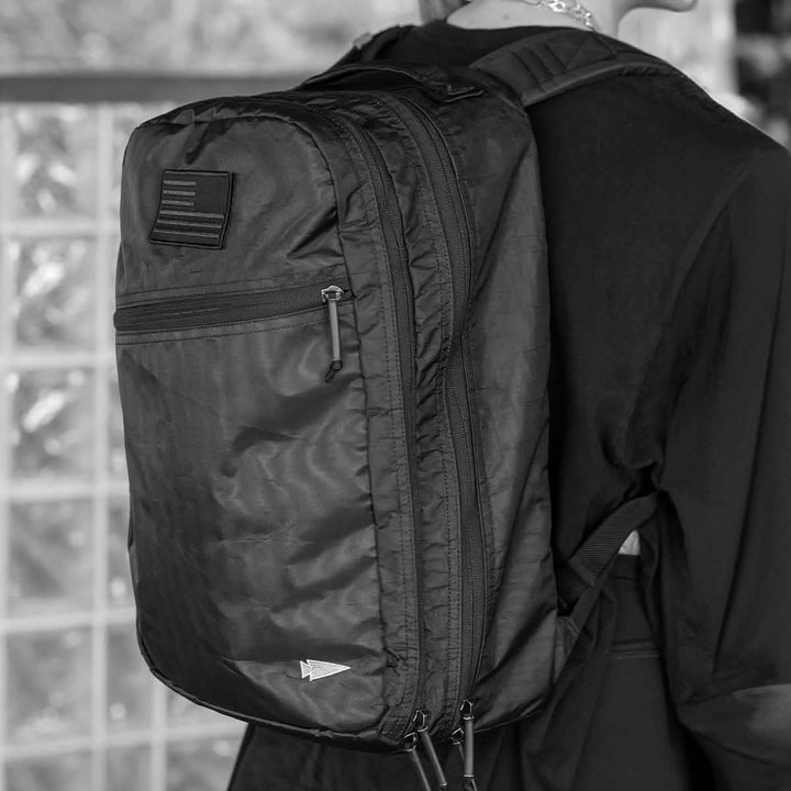 A person wearing a large black GORUCK Bullet Ruck Double Compartment - Dyneema, featuring multiple zippers and a patch on the back, stands against a wall of glass blocks. Dressed in dark clothing, they are prepared to travel hands-free with ease and style.