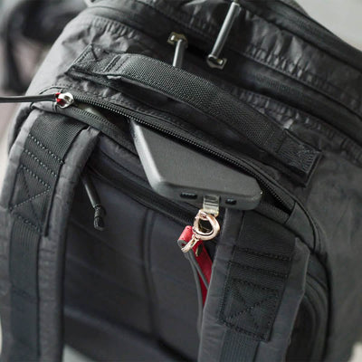 The Bullet Ruck Double Compartment - Dyneema by GORUCK is a black backpack made from robust Dyneema fabric, offering smart storage solutions. When partially unzipped, it reveals a cleverly designed hidden pocket that securely holds a smartphone. The black phone charges safely while the adjustable padded straps provide comfort. This product also comes with a lifetime guarantee.