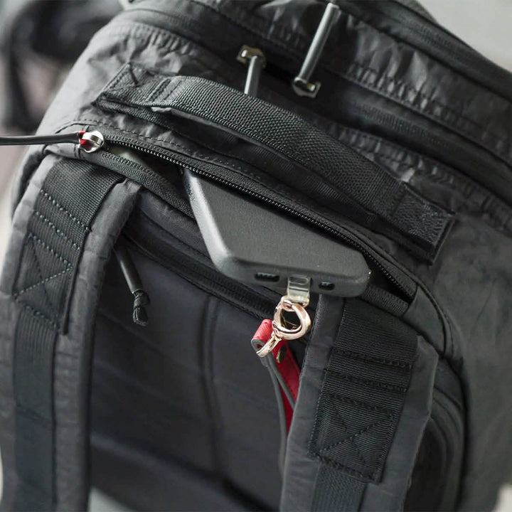 The Bullet Ruck Double Compartment - Dyneema by GORUCK is a black backpack made from robust Dyneema fabric, offering smart storage solutions. When partially unzipped, it reveals a cleverly designed hidden pocket that securely holds a smartphone. The black phone charges safely while the adjustable padded straps provide comfort. This product also comes with a lifetime guarantee.