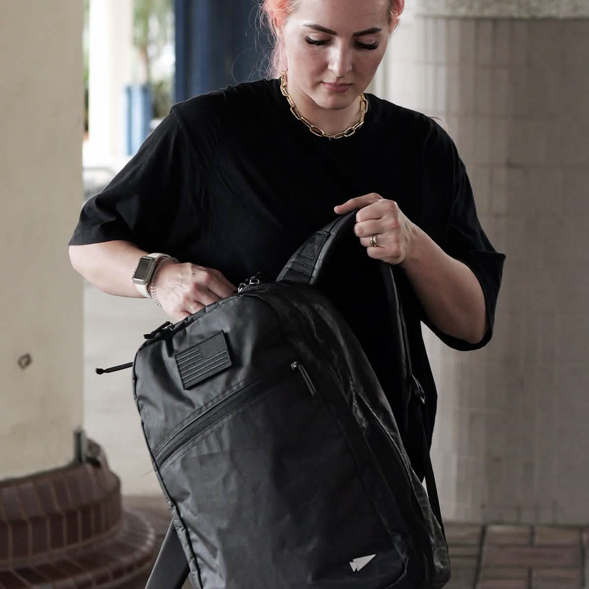 A person with pink hair wearing a black shirt and a gold chain stands under a covered area, examining their GORUCK Bullet Ruck Double Compartment backpack. Constructed from Dyneema for durability, it comes with a lifetime guarantee. They hold the bag in one hand while adjusting it with the other.