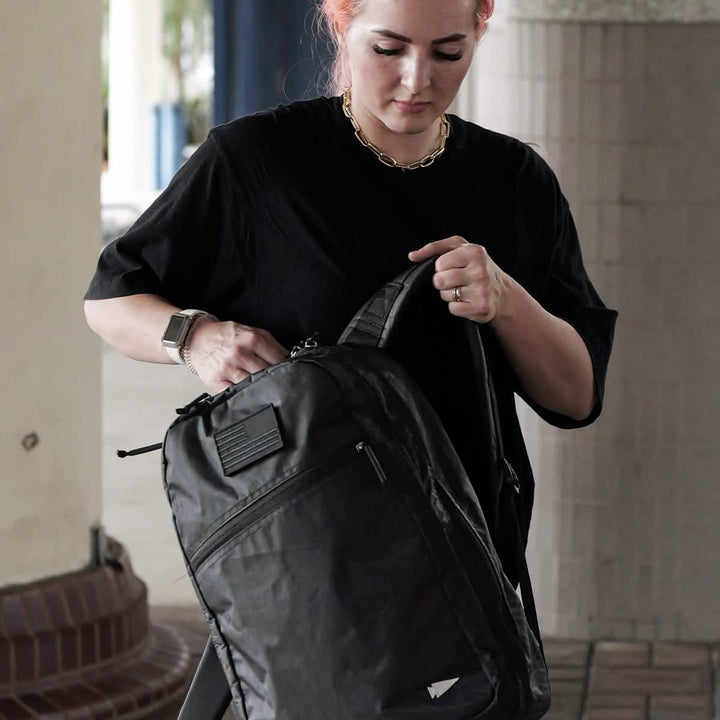 A person with pink hair wearing a black shirt and a gold chain stands under a covered area, examining their GORUCK Bullet Ruck Double Compartment backpack. Constructed from Dyneema for durability, it comes with a lifetime guarantee. They hold the bag in one hand while adjusting it with the other.