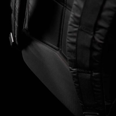 Close-up image of a black Bullet Ruck Double Compartment backpack by GORUCK, emphasizing the shoulder strap area with detailed stitching and a padded section. The dark backdrop accentuates the texture and design of the durable Dyneema fabric, which is backed by a lifetime guarantee.