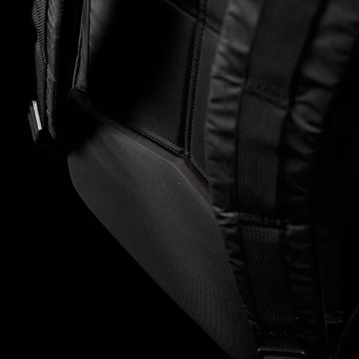Close-up image of a black Bullet Ruck Double Compartment backpack by GORUCK, emphasizing the shoulder strap area with detailed stitching and a padded section. The dark backdrop accentuates the texture and design of the durable Dyneema fabric, which is backed by a lifetime guarantee.