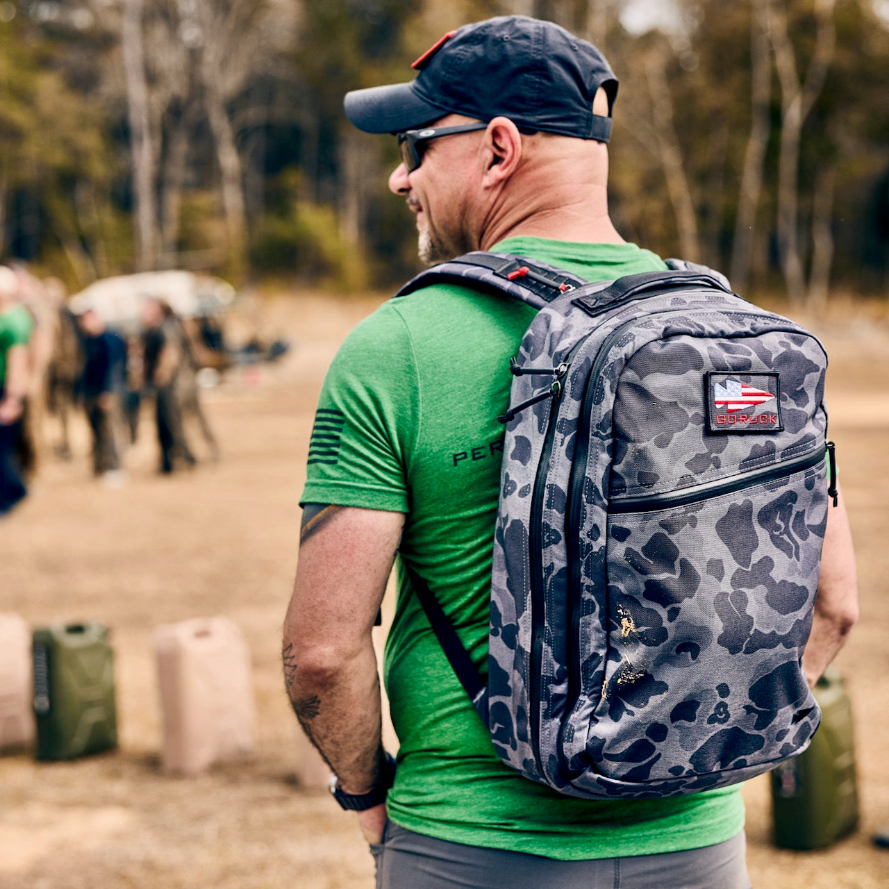 Man in a green shirt and cap with the Double Compartment Bullet Ruck - Ballistic Nylon Cordura - 18L, featuring a bombproof laptop compartment, stands outdoors among people and gear.