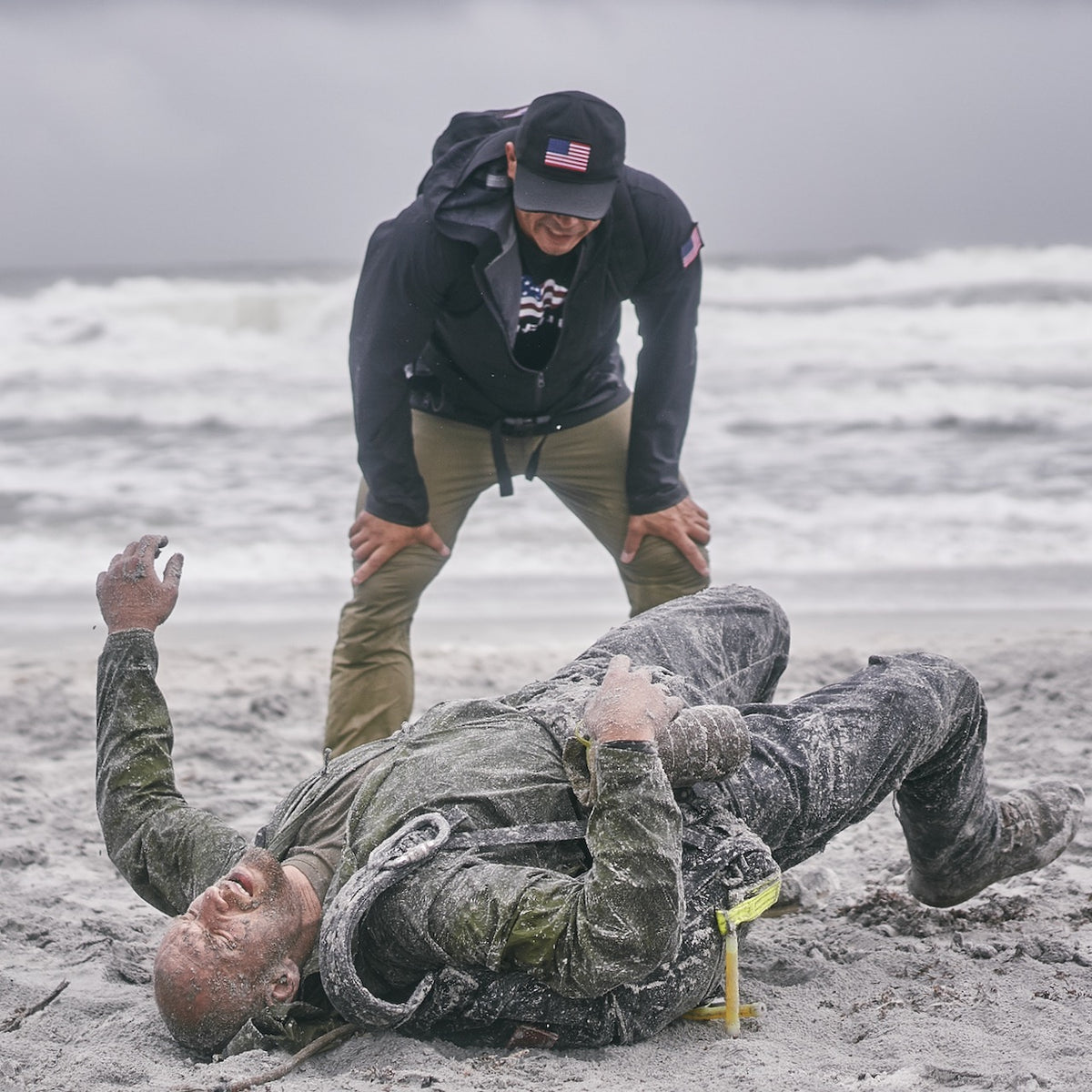 A man lies on a sandy beach, covered in sand, as another man wearing a Cadre Hat - CORDURA® stands over him, both near the ocean.