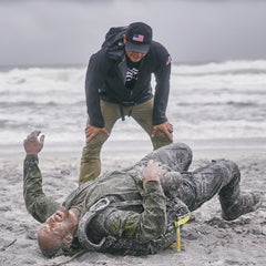 A man lies on a sandy beach, covered in sand, as another man wearing a Cadre Hat - CORDURA® stands over him, both near the ocean.