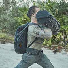 Wearing the Padded Hip Belt, a person punches a sandbag outdoors amidst trees and plants on a dirt path, maximizing the benefits of their rucking workout.