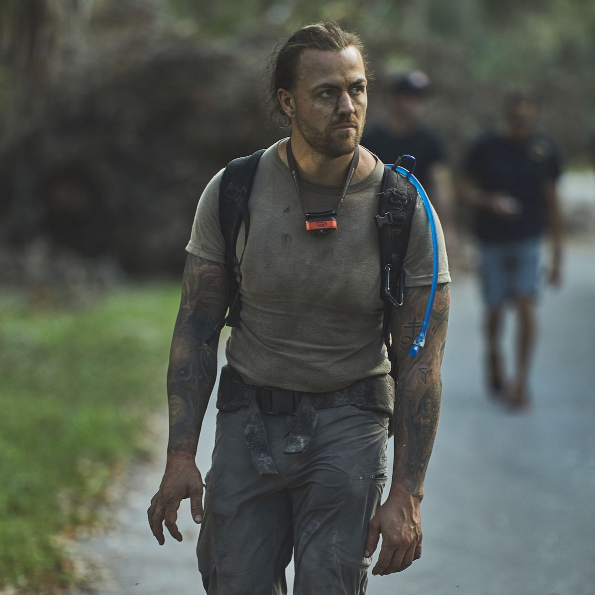 A man with tattoos and a hydration pack walks outdoors, looking serious, his gear featuring the Padded Hip Belt for added comfort during rucking. Another person is blurred in the background.