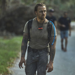 A man with tattoos and a hydration pack walks outdoors, looking serious, his gear featuring the Padded Hip Belt for added comfort during rucking. Another person is blurred in the background.