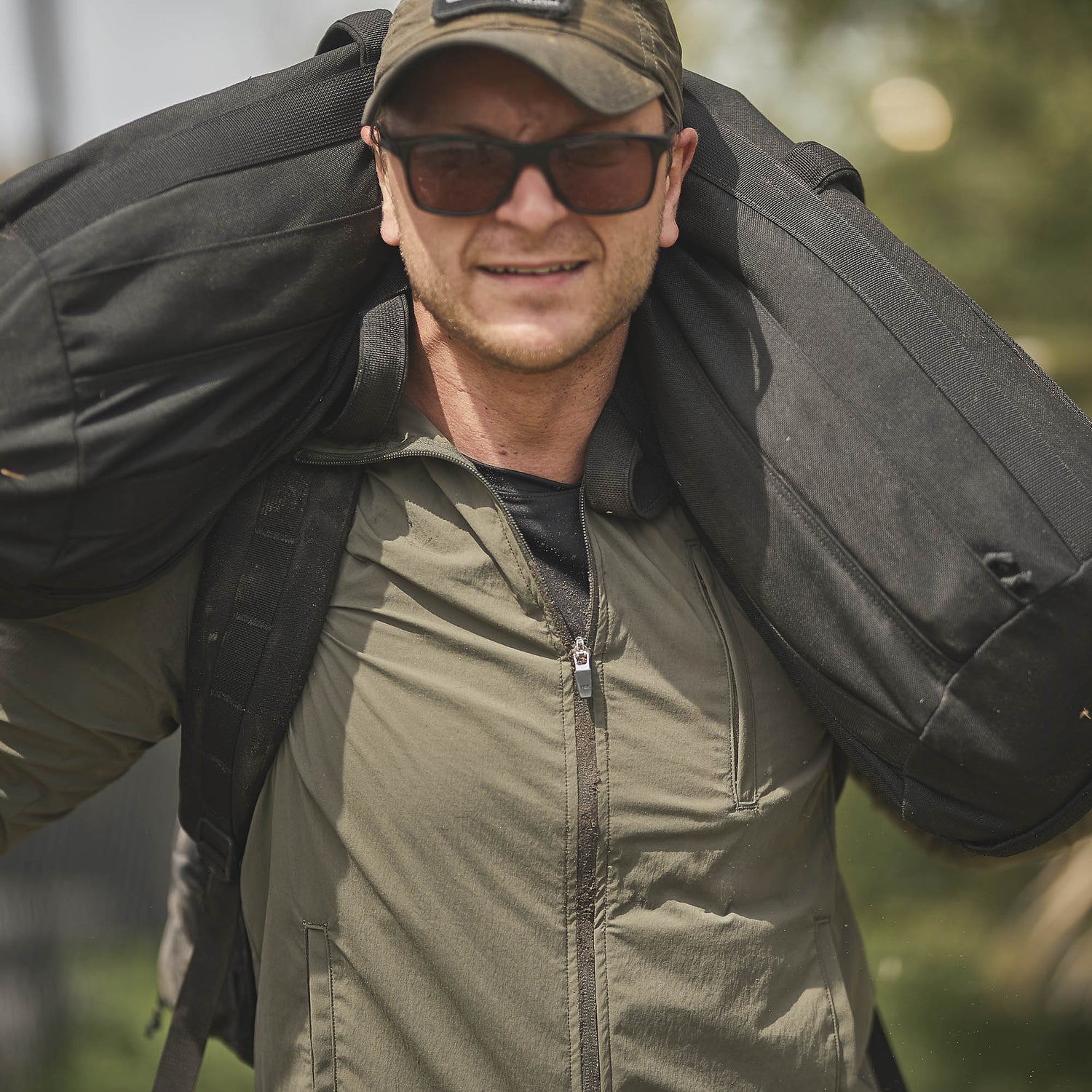 Man in sunglasses and a cap carrying a large black bag on his shoulders, wearing a green Indestructible Challenge Windbreaker jacket.